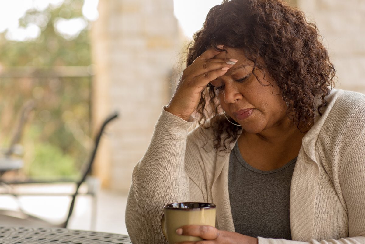 woman holds forehead while sitting at a table with coffee mug