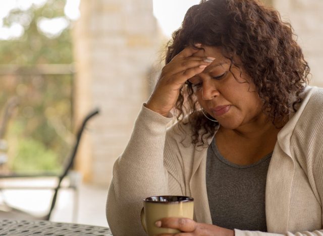 woman holds forehead while sitting at a table with coffee mug