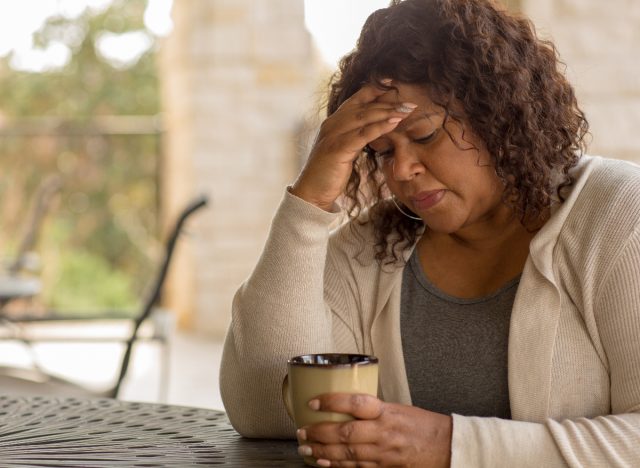 woman holds forehead while sitting at a table with coffee mug