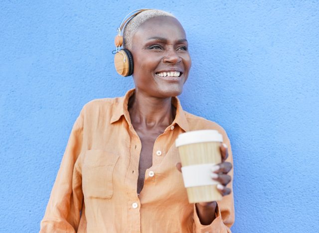 happy woman holds coffee cup while listening to music on headphones, standing in front of blue wall