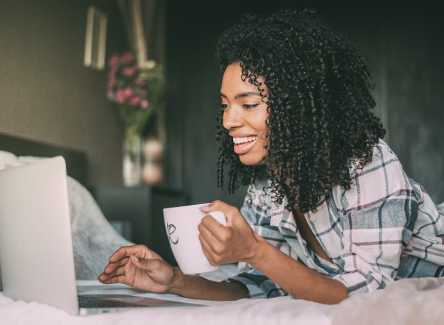 happy woman laying on bed with mug of tea on laptop