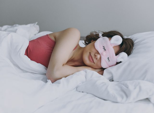mulher se aconchega na cama de pelúcia usando máscara de dormir rosa