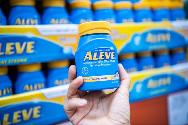 Shoppers hand holding a plastic container of Bayer's Aleve brand genuine noxopren sodium tablets in supermarket aisle.