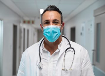 Portrait of mixed race male doctor wearing face mask standing in hospital corridor.