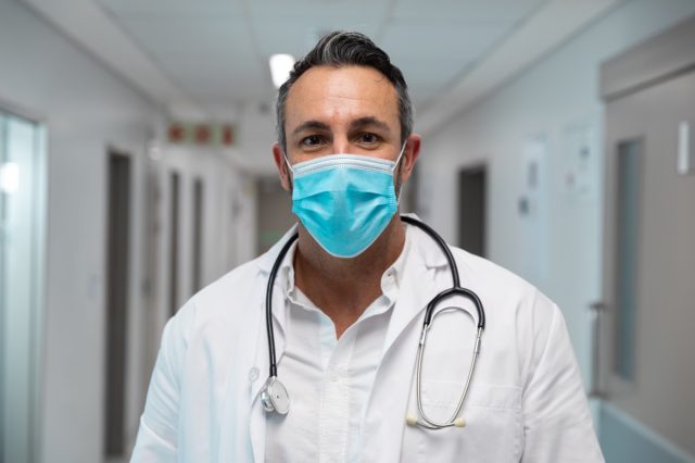 Portrait of mixed race male doctor wearing face mask standing in hospital corridor.