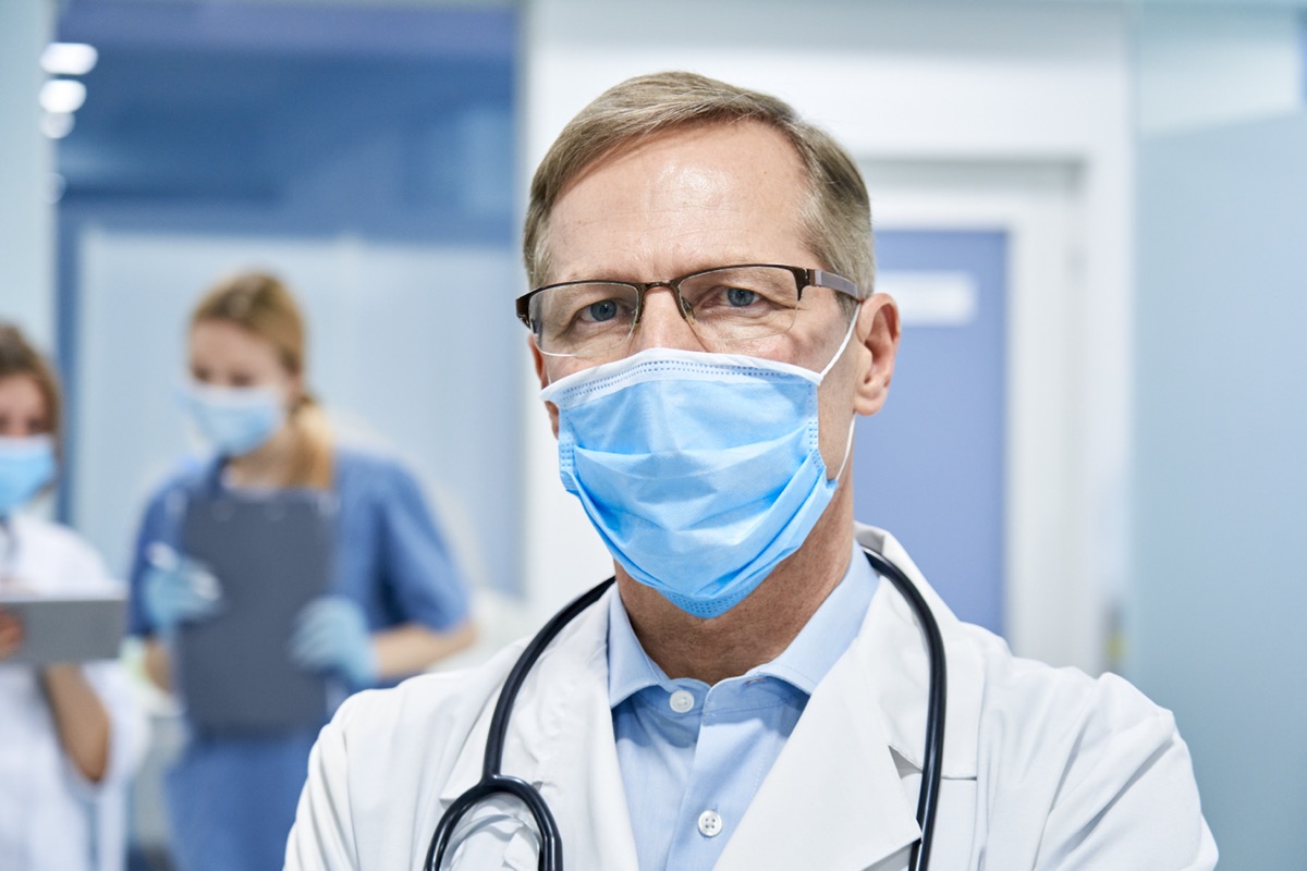 Mature old medical healthcare professional doctor wearing white coat, stethoscope, glasses and face mask standing in hospita.l looking at camera