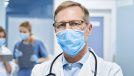 Mature old medical healthcare professional doctor wearing white coat, stethoscope, glasses and face mask standing in hospita.l looking at camera