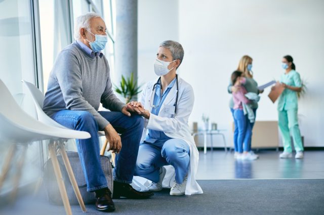 Verstörter älterer Mann sitzt im Wartezimmer des Krankenhauses, während die Ärztin seine Hand hält und ihn tröstet