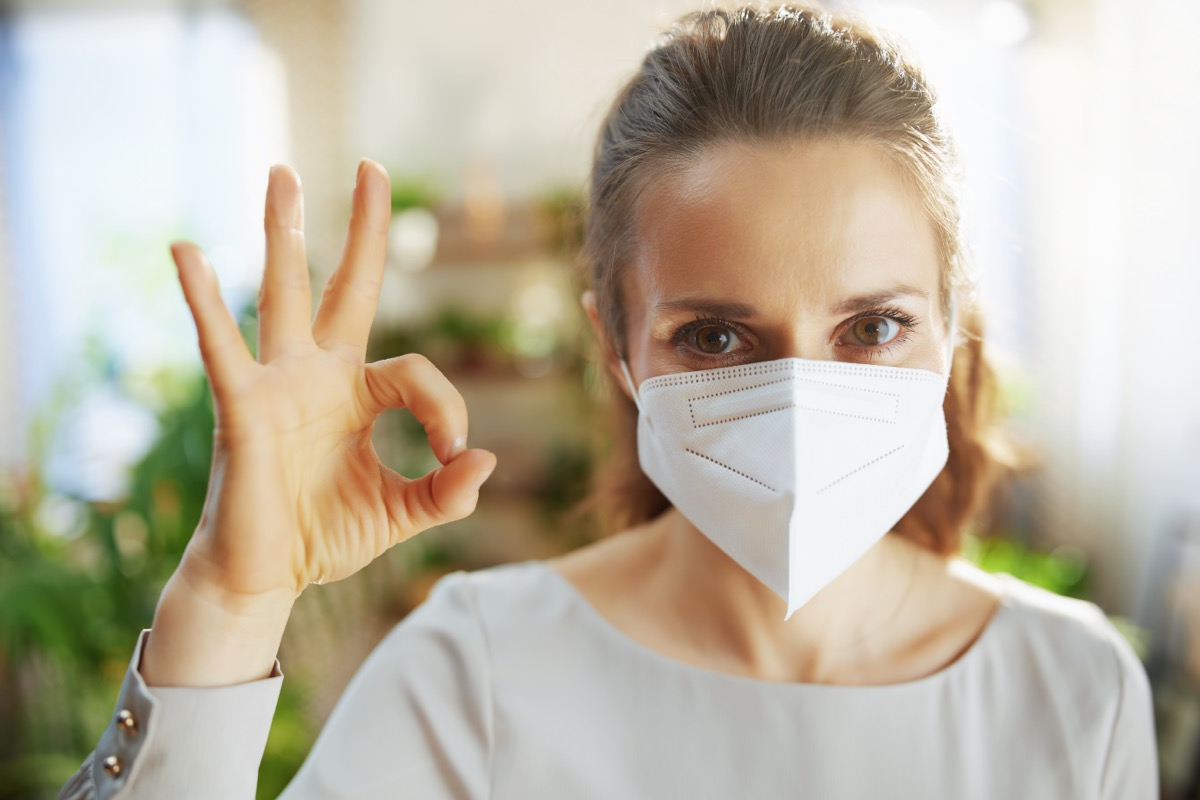Smiling middle aged woman in grey blouse with ffp2 mask at modern home showing ok gesture.