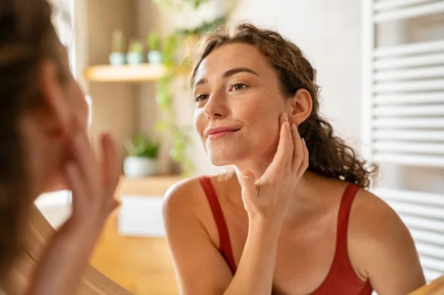 Young woman looking at mirror while touching her face.