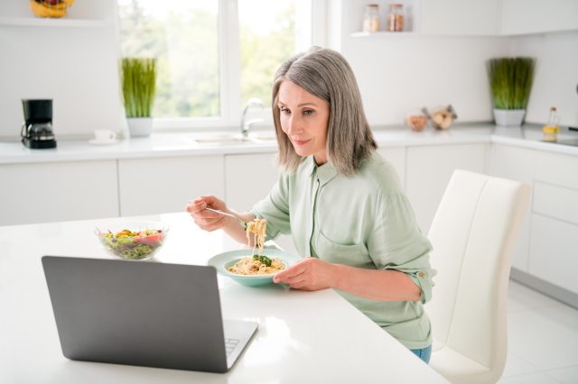 Portrait of attractive focused grey-haired woman eating homemade dinner watching video at home light white indoors