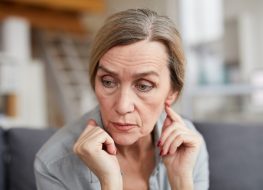 Portrait of sad mature woman sitting on couch at home and looking away with worry and anxiety.