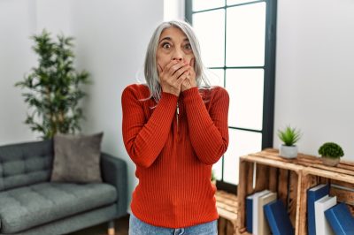 Middle age grey-haired woman wearing casual clothes standing at home shocked covering mouth with hands.