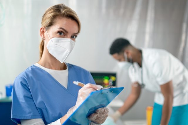 Portrait of a doctor with a face mask and a clipboard looking at a camera in a hospital.