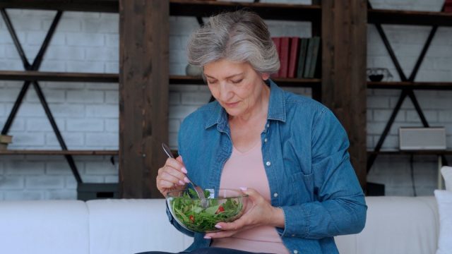 Elderly woman eats her vegetable salad sitting on the sofa at home.