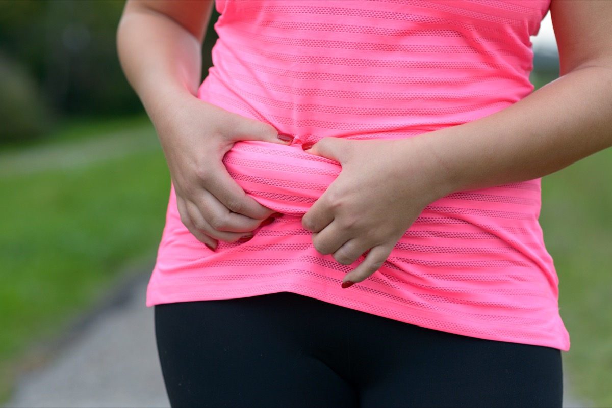 Woman wearing a pink shirt outdoors holding a roll of excess fat around her stomach area pinched in her hands in a close up torso view.