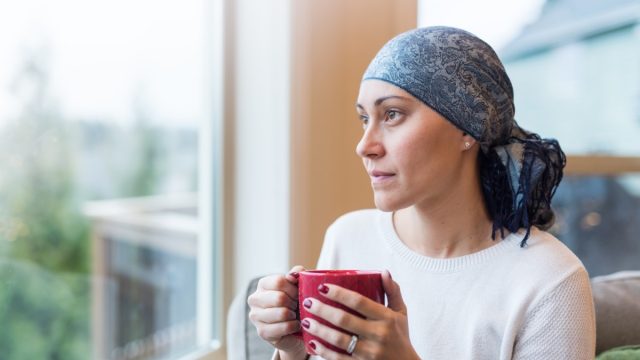 Woman in her 30s sits by her living room window with a cup of tea and looks out contemplatively. She is a cancer survivor and is wearing a headscarf.
