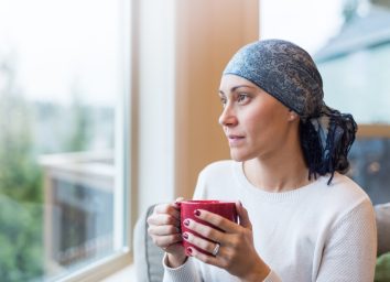 Woman in her 30s sits by her living room window with a cup of tea and looks out contemplatively. She is a cancer survivor and is wearing a headscarf.