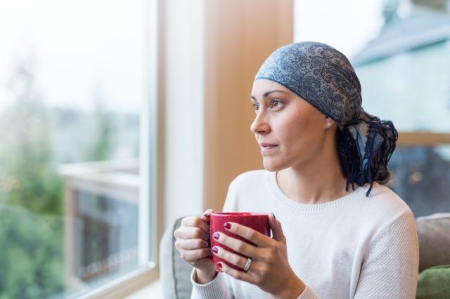 Woman in her 30s sits by her living room window with a cup of tea and looks out contemplatively. She is a cancer survivor and is wearing a headscarf.