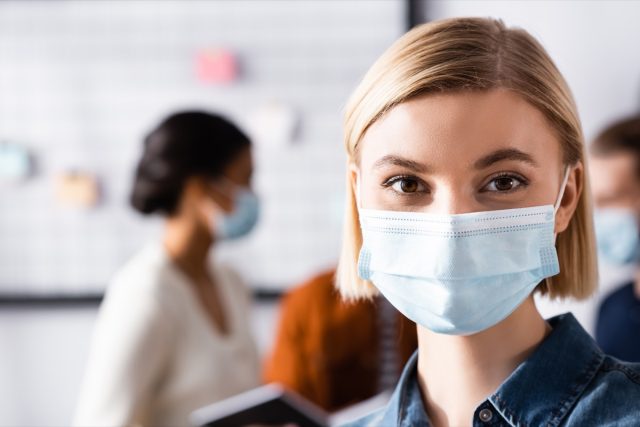 Blonde businesswoman in medical mask looking at camera while multiethnic colleagues talking on blurred background.