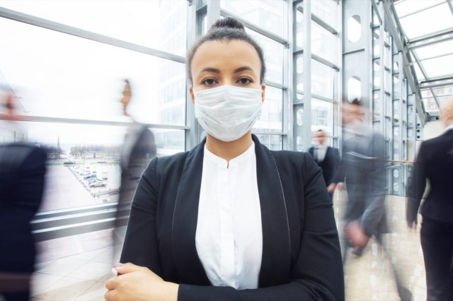 Business woman in suit wearing surgical protect mask standing in a crowd of walking people.