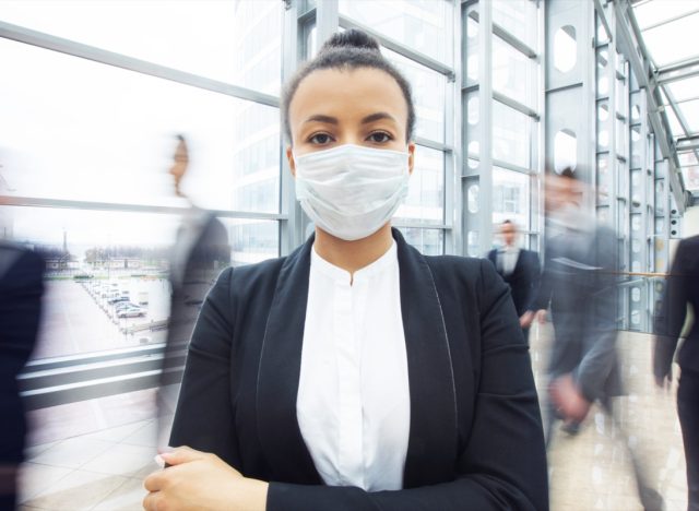 Business woman in suit wearing surgical protect mask standing in a crowd of walking people.