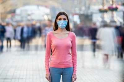 The young girl with medical mask on her face stands on the crowded street.