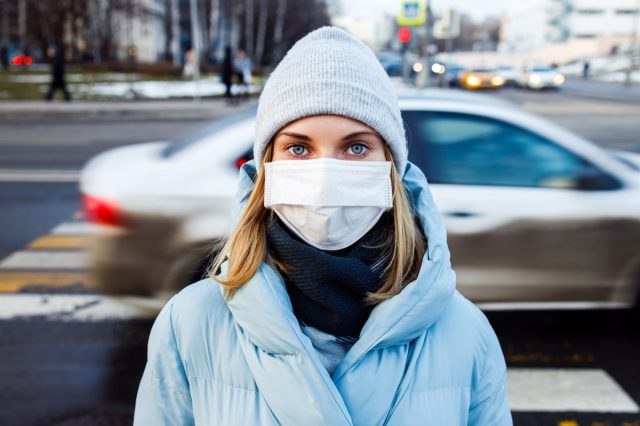 Young woman in mask looking at camera on walk on street in city during day.