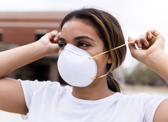 A mid adult woman protects herself by placing an N95 face mask over her nose and mouth.