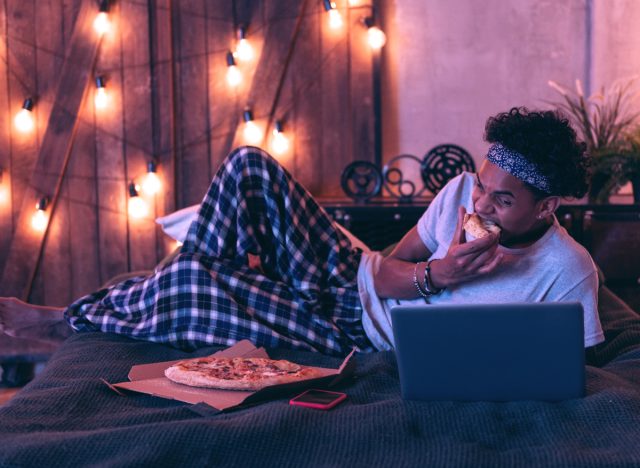 man lounging in bed in PJs while eating pizza