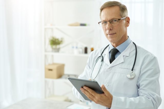 Handsome doctor in lab coat using tablet computer in clinic.