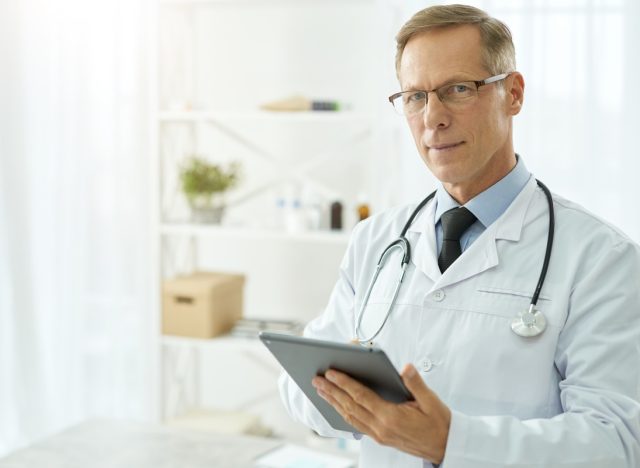 Handsome doctor in lab coat using tablet computer in clinic.