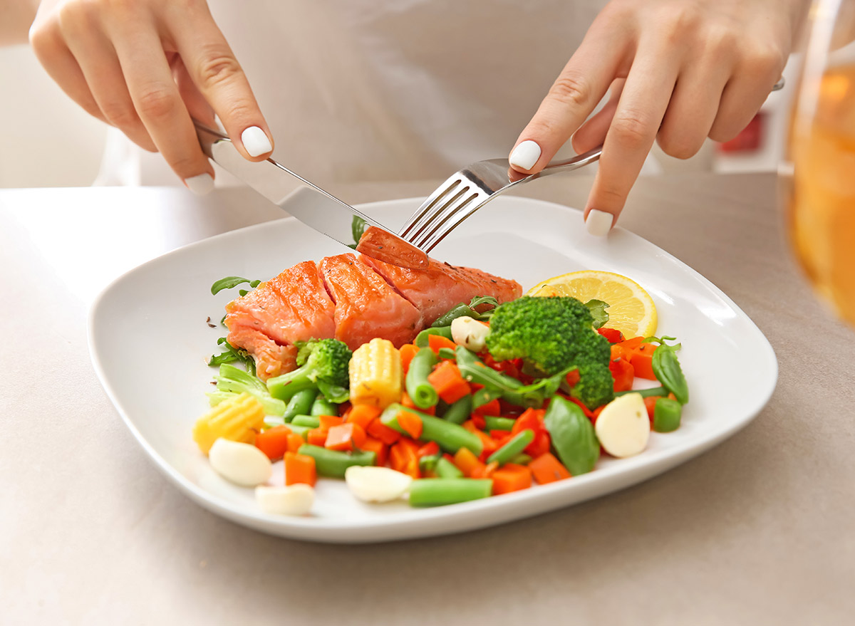 woman cutting into salmon with plate of veggies