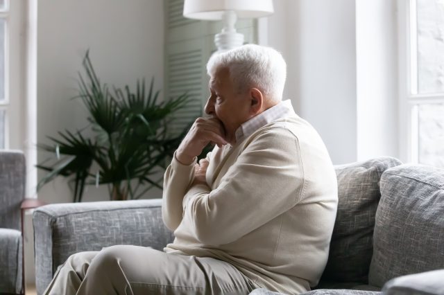 Grey haired senior male seats on couch in living room.