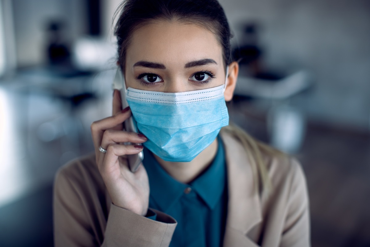 Close-up of a businesswoman with face mask making a phone call over mobile phone while working in the office and looking at camera.