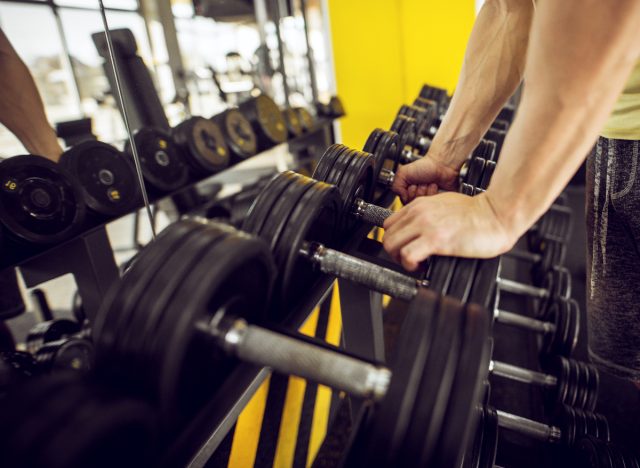 close-up man's arms grabbing dumbbells in gym mirror