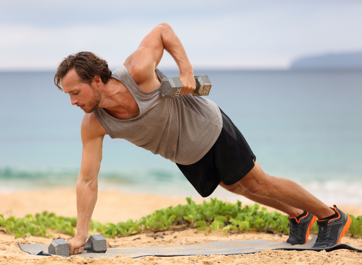man doing renegade row with dumbbells on mat outside by beach