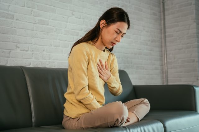 A young Asian woman feels uncomfortable with heartburn while holding her chest with eyes closed and sitting with legs folded on the sofa at home.