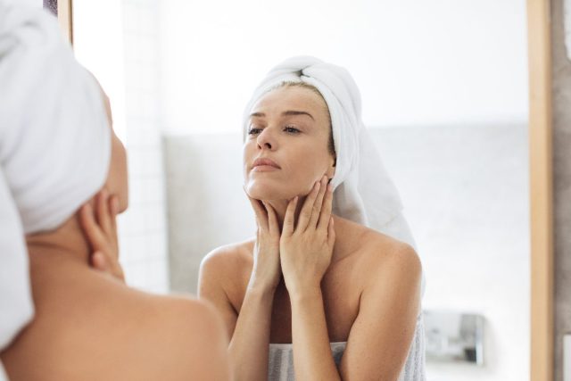 woman touching her neck, skincare, looking in bathroom mirror