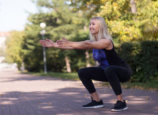 Žena středního věku dřepí s tělesnou hmotností venku při chůzi