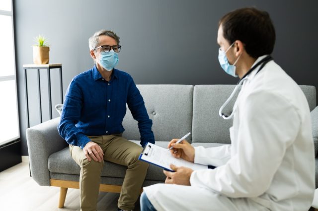 Doctor With Senior Patient Living At Home In Mask.