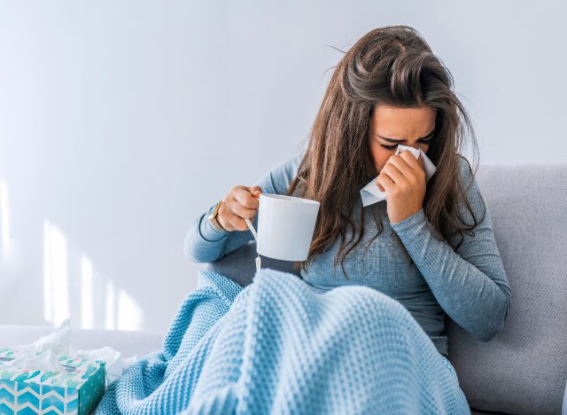 woman with seasonal allergies blowing nose on couch under blanket