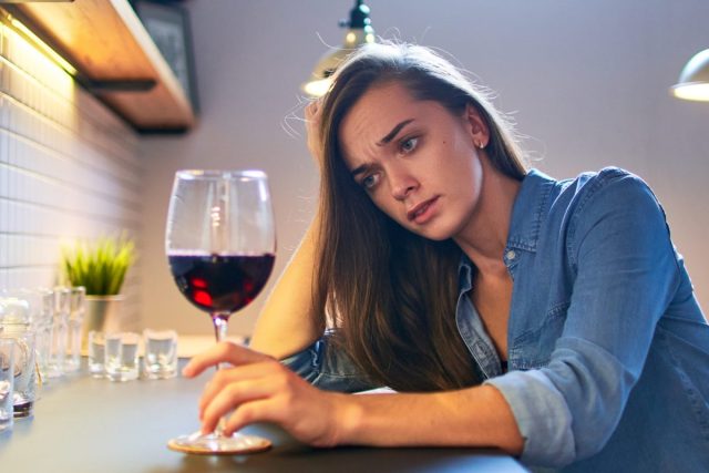 A sad woman drinking wine in the kitchen.