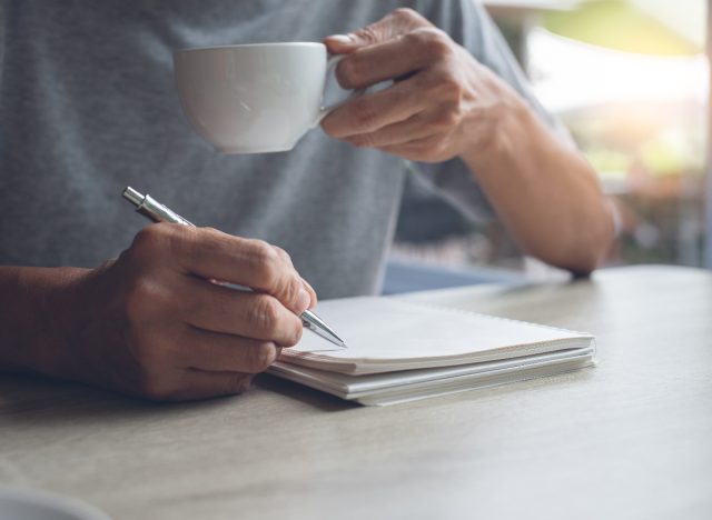 man holding coffee cup and writing in notebook