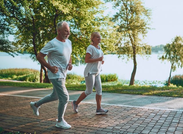 mature couple running outdoors by lake, demonstrating cardio habits that are aging you faster