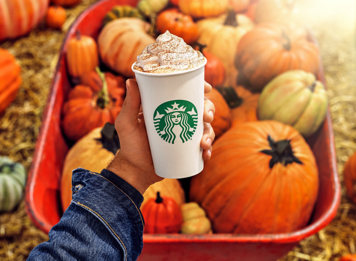Person holding Starbucks' Pumpkin Spice Latte in front of wheelbarrow of pumpkins