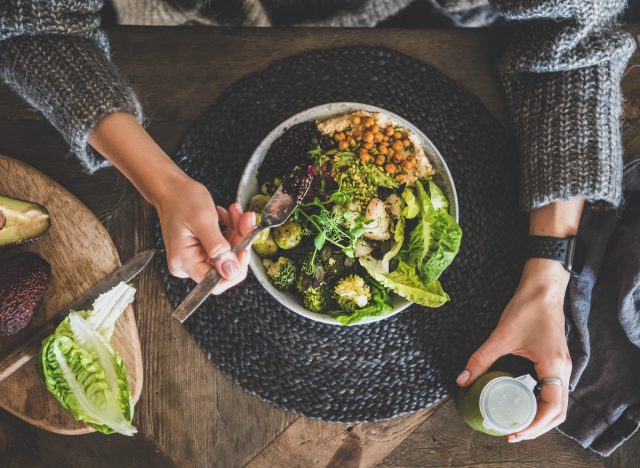 woman holding a smoothie and eating a grain bowl with chickpeas, vegetables, hummus, and couscous