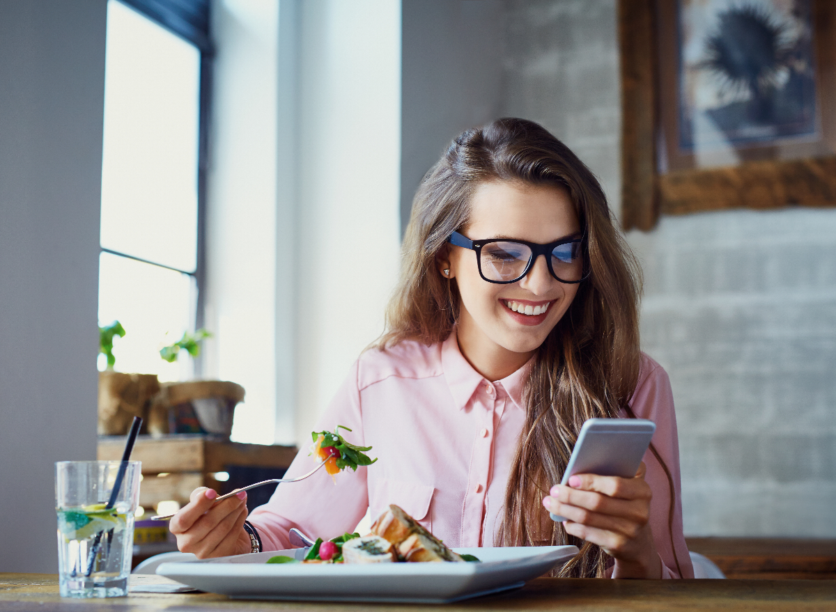 woman eating salad at restaurant while looking at phone