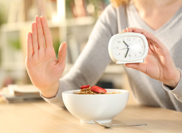 woman holding up hand and clock showing intermittent fasting concept with bowl of cereal