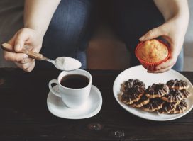 woman pouring sugar into coffee, holding muffin above plate of cookies